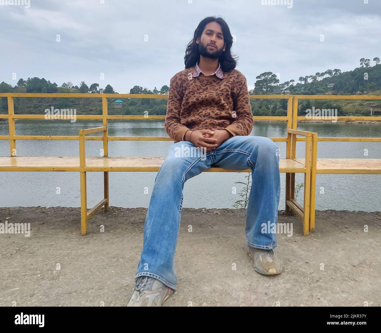 Vorderansicht eines gut aussehenden indischen jungen Mannes mit langen Haaren und Bart, der auf die Kamera schaute, während er auf der Bank neben dem See saß Stockfoto