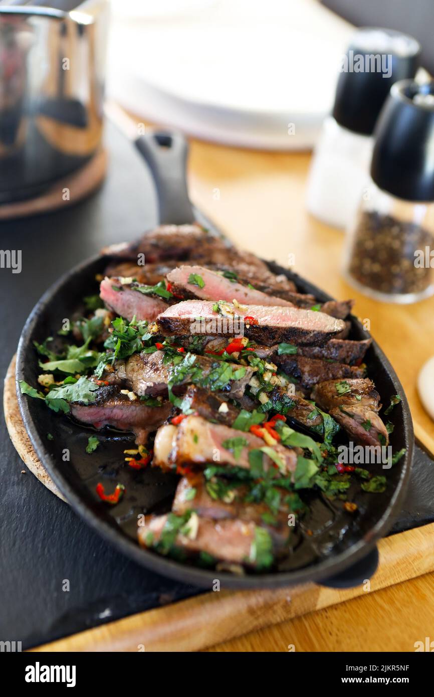 Ribeye Steak in Scheiben mit Koriander, Knoblauch und Chilis in einer heißen Pfanne auf dem Esstisch, fertig zum Servieren Stockfoto