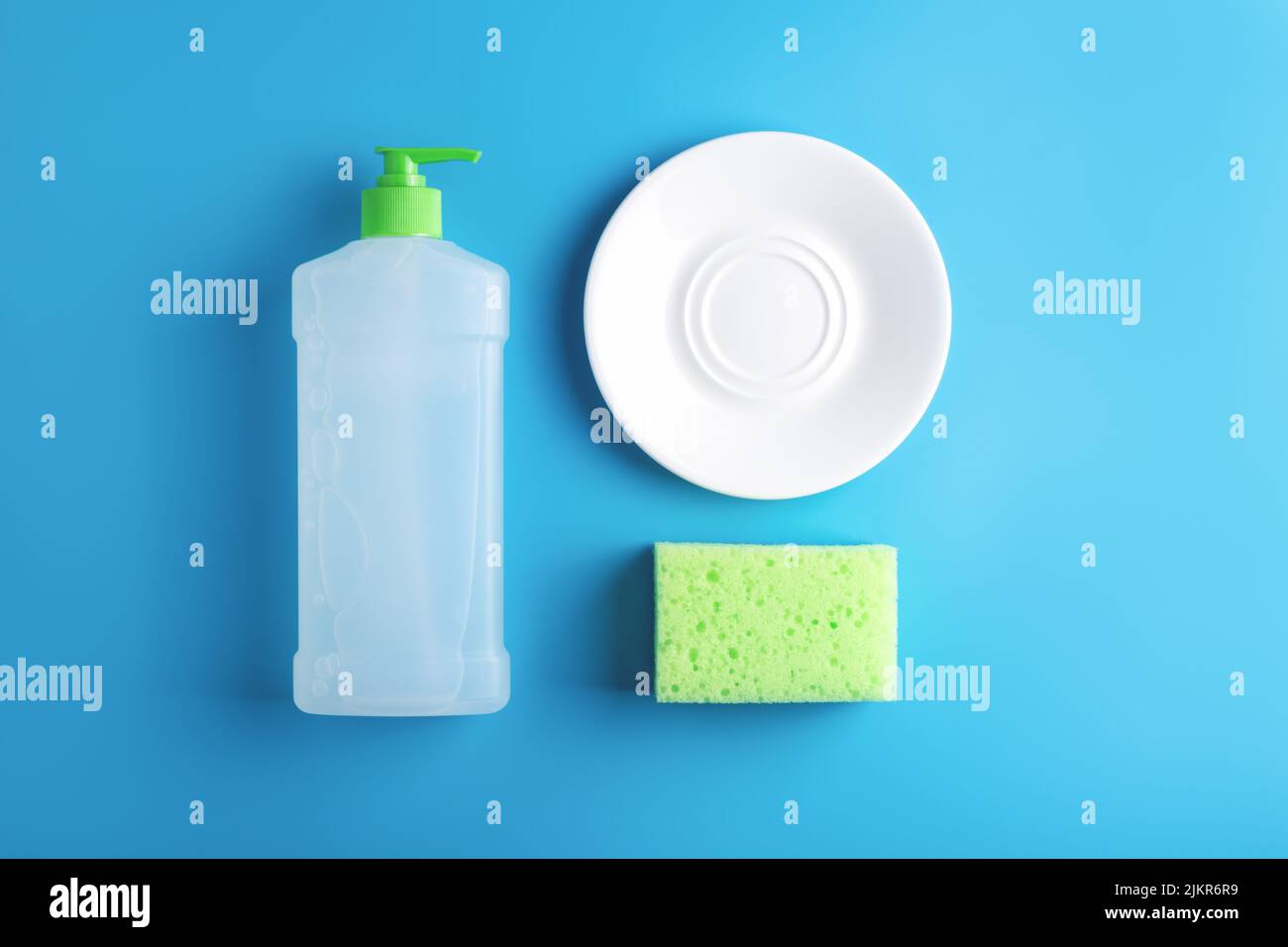 Küchenzusammensetzung mit sauberem Glas weißen Teller, grünen Reinigungsschwamm und Flasche Spülmittel auf blauem Hintergrund angeordnet. Flach liegend, Haushaltskerne. Stockfoto