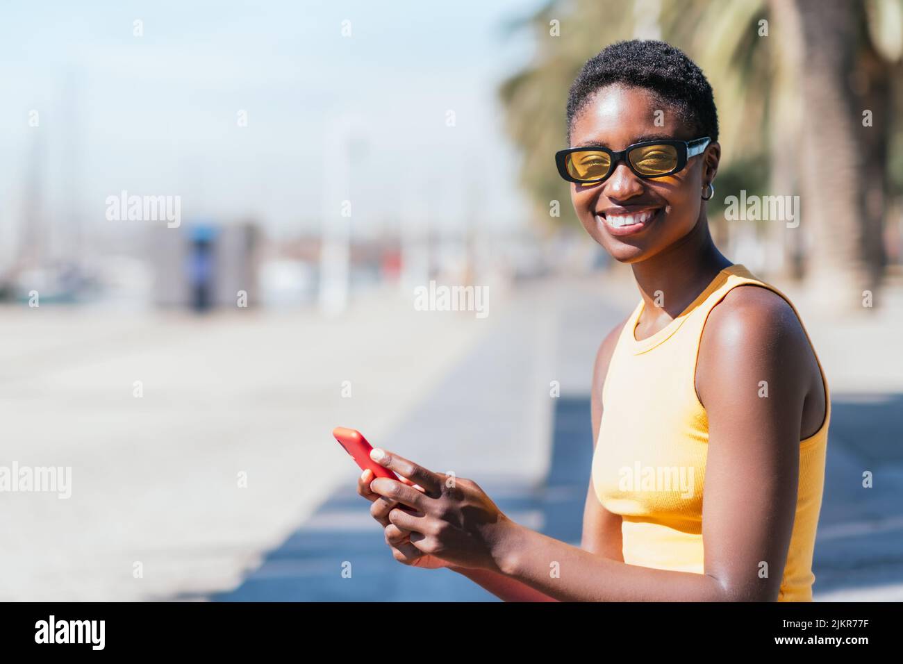 Positive afrikanische Frau, die die Kamera anschaut, während sie ein rotes Smartphone hält. Sie hat einen trendigen kurzen Haarschnitt und eine lässige Sommerkutte Stockfoto