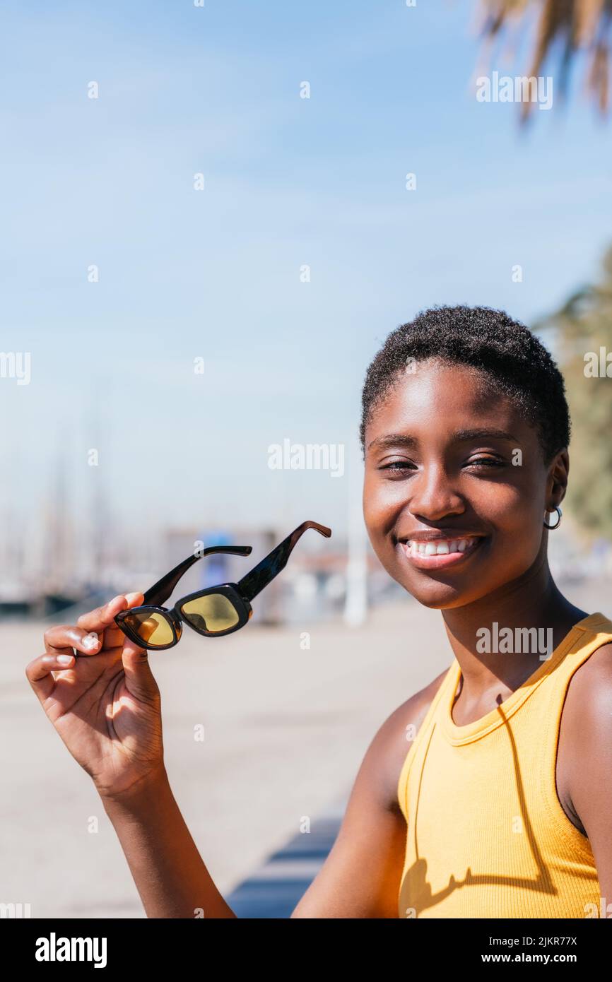 Porträt einer positiven afrikanischen Frau, die in Küstennähe auf die Kamera schaut. Sie trägt ein sommerliches gelbes T-Shirt und trägt eine gelbe Sonnenbrille Stockfoto