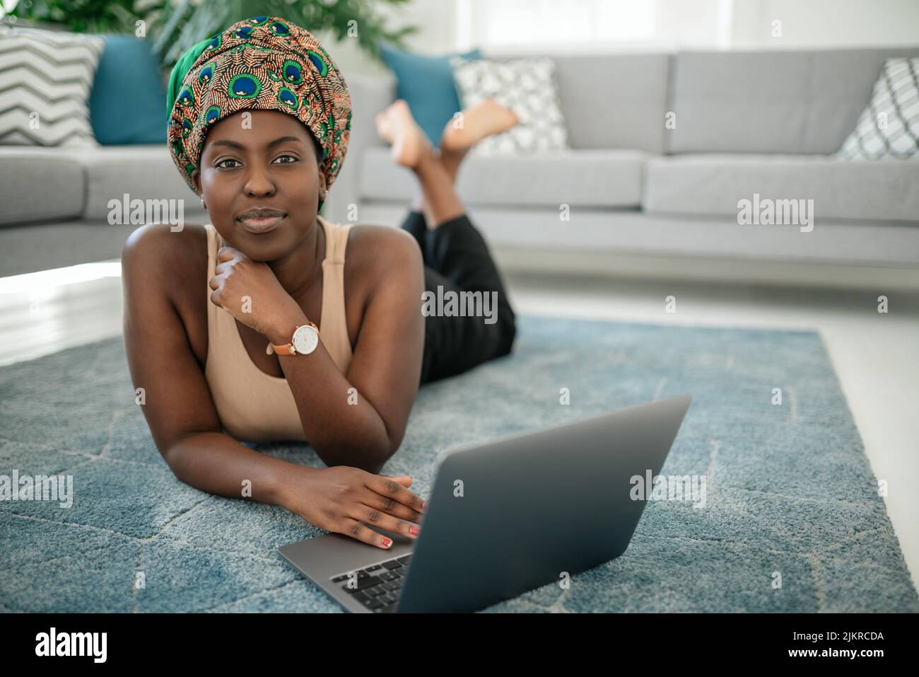 Glückliche junge afrikanische Frau, die zu Hause mit einem Laptop die Beine gekreuzt auf ihrem Teppich sitzt und in die Kamera schaut Stockfoto