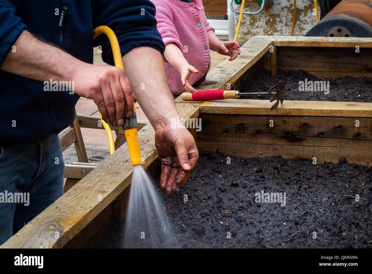 Vater und Tochter Garten zusammen. Der Mensch sprüht Wasser auf frisch gepflanzte Samen. Mädchen mit Gartenwerkzeugen hilft. Stockfoto