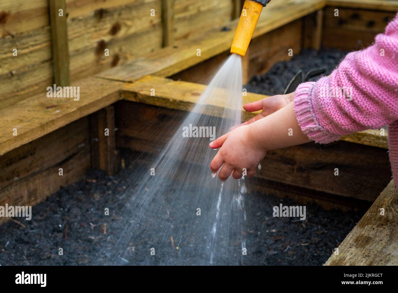 Kleines Kind hält die Hände unter Wasser-Schlauchspray an der Gemüsekiste im Garten. Stockfoto
