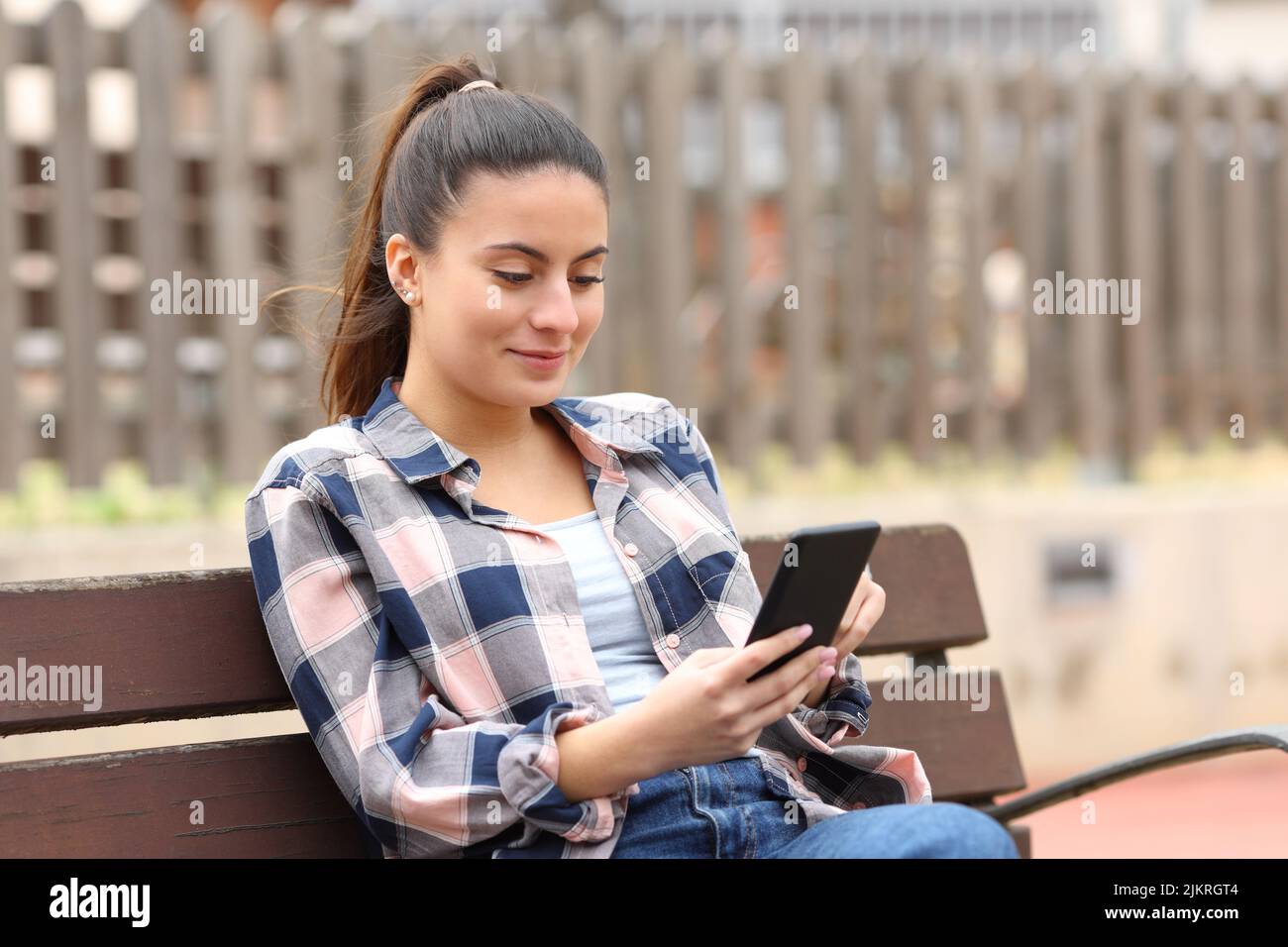 Glücklicher Teenager, der ein Handy benutzt und auf einer Bank in einem Park sitzt Stockfoto