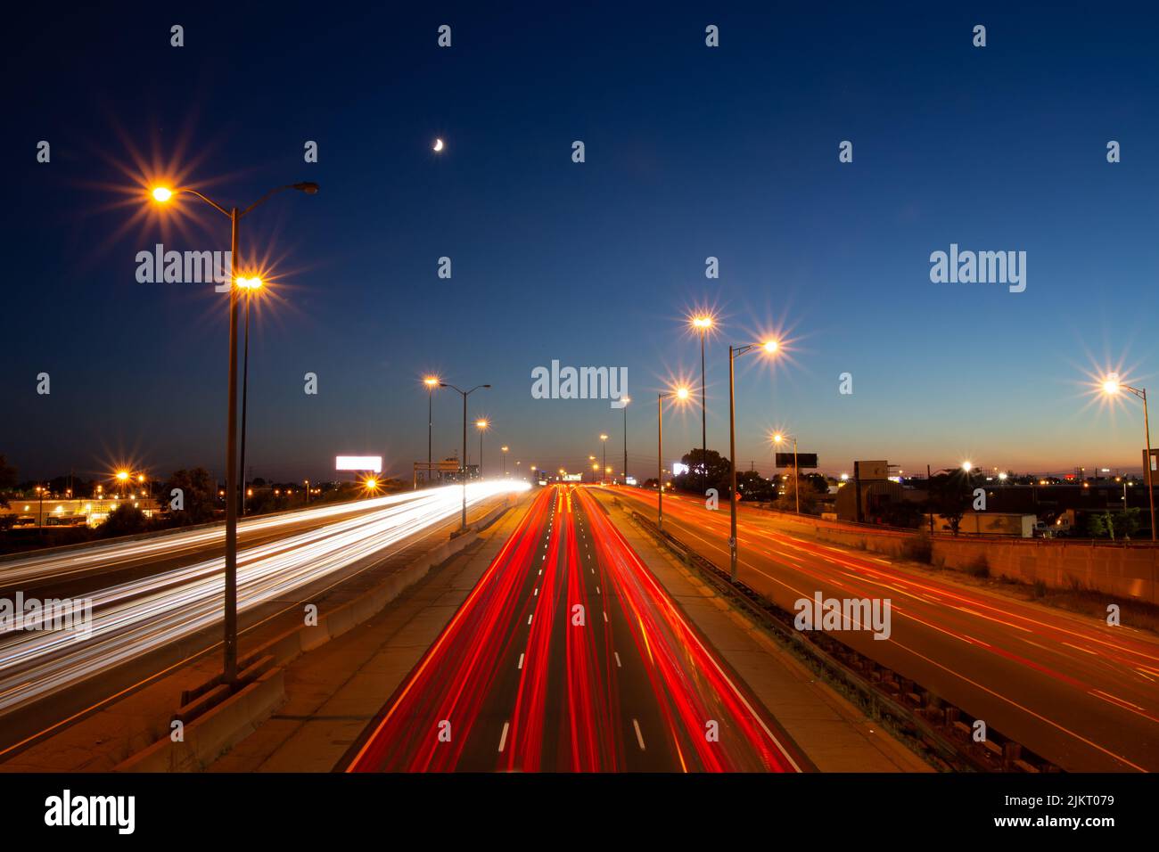 Langzeitaufnahme des geschäftigen Verkehrs auf einer Autobahn unter dem Mond und dem blauen und orangenen Sonnenuntergangshimmel in Toronto, Ontario, Kanada. Stockfoto