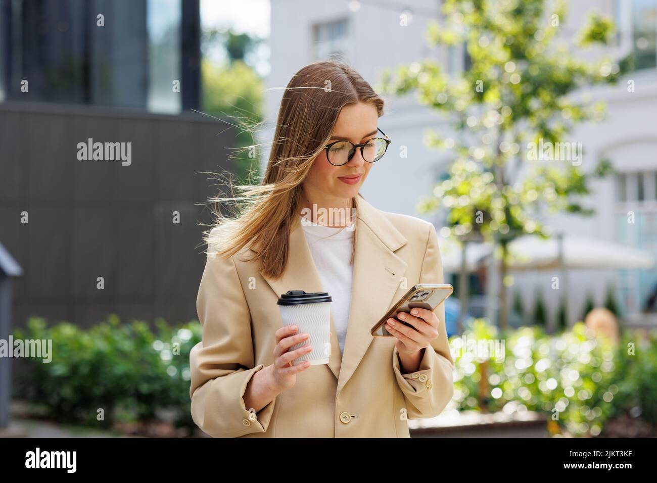 Ein junges blondes Mädchen vor dem Hintergrund eines Geschäftszentrums, Bürozentrums. Porträt eines erfolgreichen Start-ups. Freizeitanzug mit Smartphone und Kaffee. Stockfoto