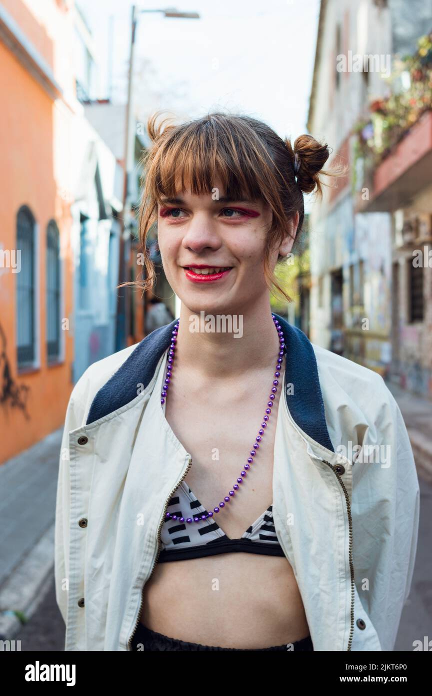 Die junge kaukasische lateinische Transsexuelle, mit rotem Make-up und leicht bekleidet, steht in einer städtischen Gasse und lächelt die Kamera zur Seite Stockfoto