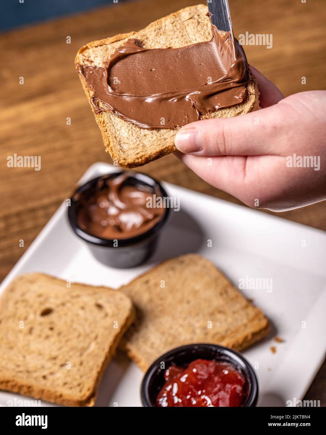 Eine Person, die Schokolade auf einem Toast verteilt, und ein Teller mit Marmelade, Schokolade und Toast auf einem Holztisch Stockfoto
