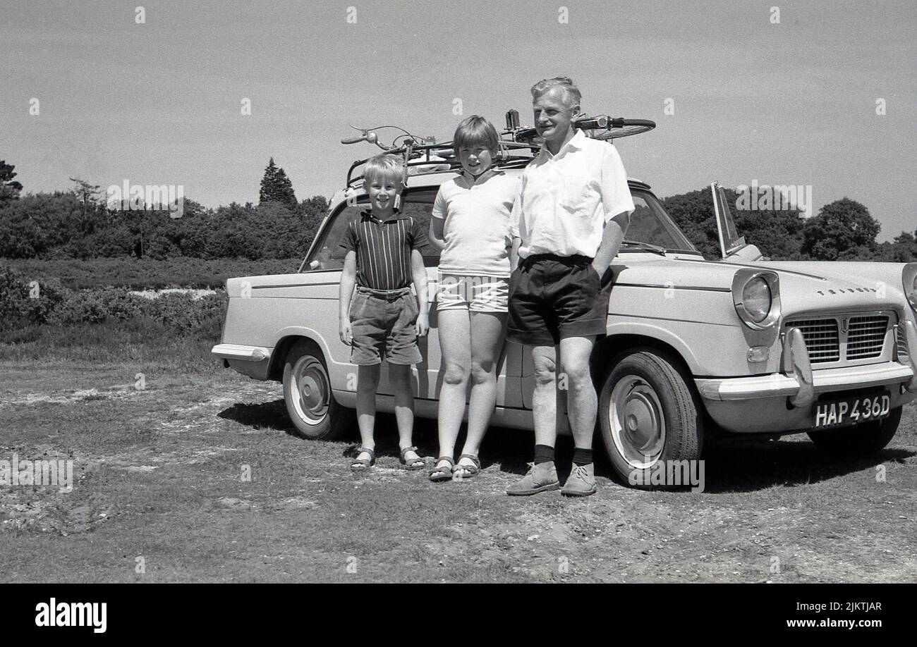 1960s, historisch, ein Vater mit seinen zwei kleinen Kindern, die neben ihrem Triumph Herald Auto der Ära stehen. Eine Fahrradwohnung auf dem Dachgepäckträger. Zwischen 1959 und 1971 von Standard-Triumph aus Coventry, England, hergestellt, war der Herald ein kleiner zweitüriger Wagen, der vom Italiener Giovanni Michelotti entworfen wurde. Stockfoto