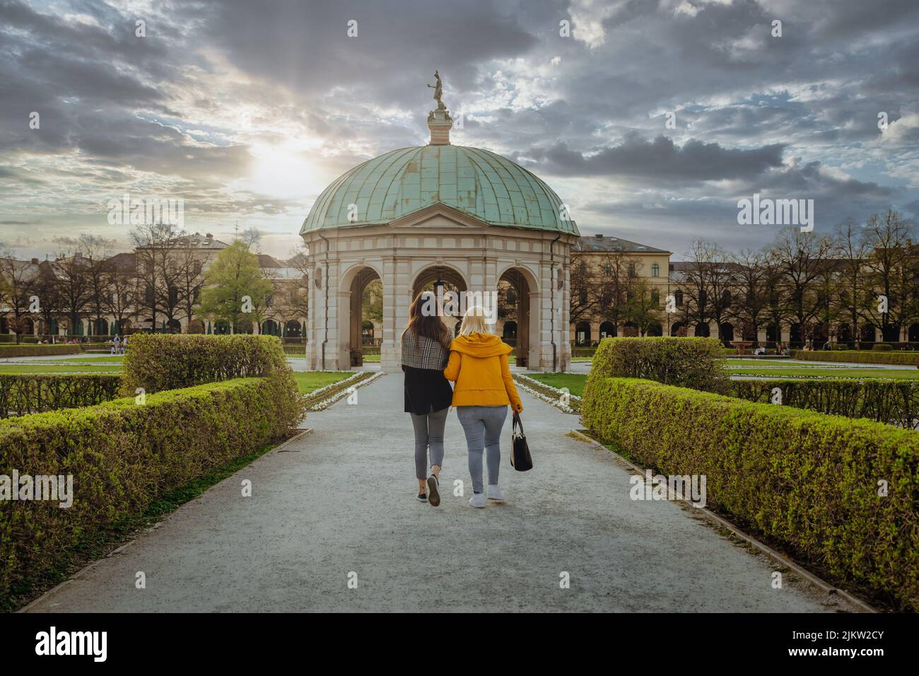 Zwei junge Freunde gehen durch den Hofgarten, den Park in der Münchner Innenstadt. Episch atemberaubend schöner Sonnenuntergang im Hintergrund des Stadtparks Stockfoto