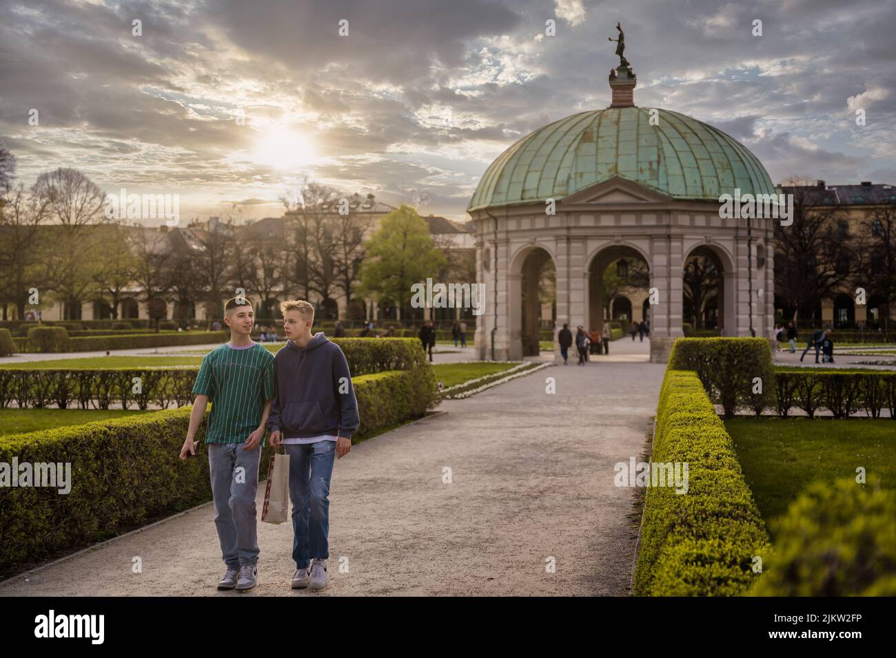 Zwei junge Freunde gehen durch den Hofgarten, den Park in der Münchner Innenstadt. Episch atemberaubend schöner Sonnenuntergang im Hintergrund des Stadtparks Stockfoto