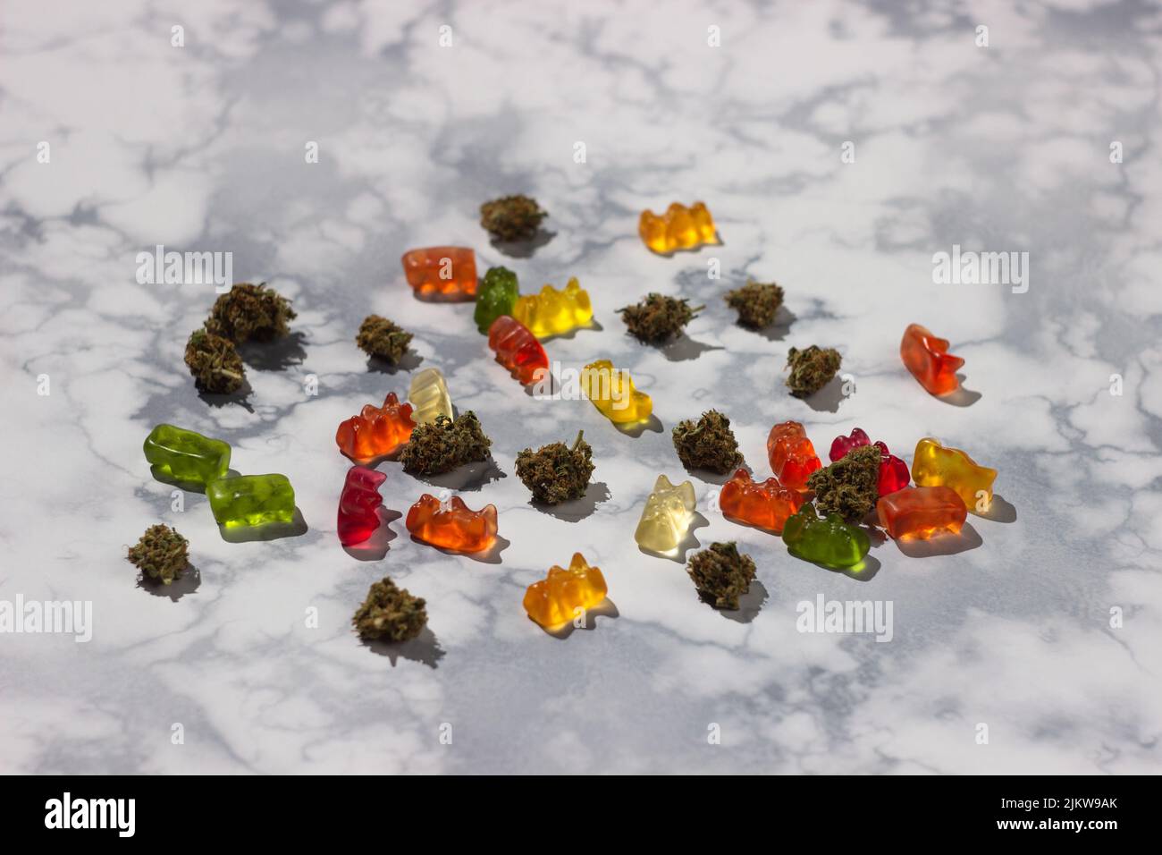 Mehrere trockene Knospen von medizinischem Marihuana liegen zufällig unter Gummibären verschiedener Farben mit der Zugabe von cbd-Öl auf einem Marmortisch Stockfoto