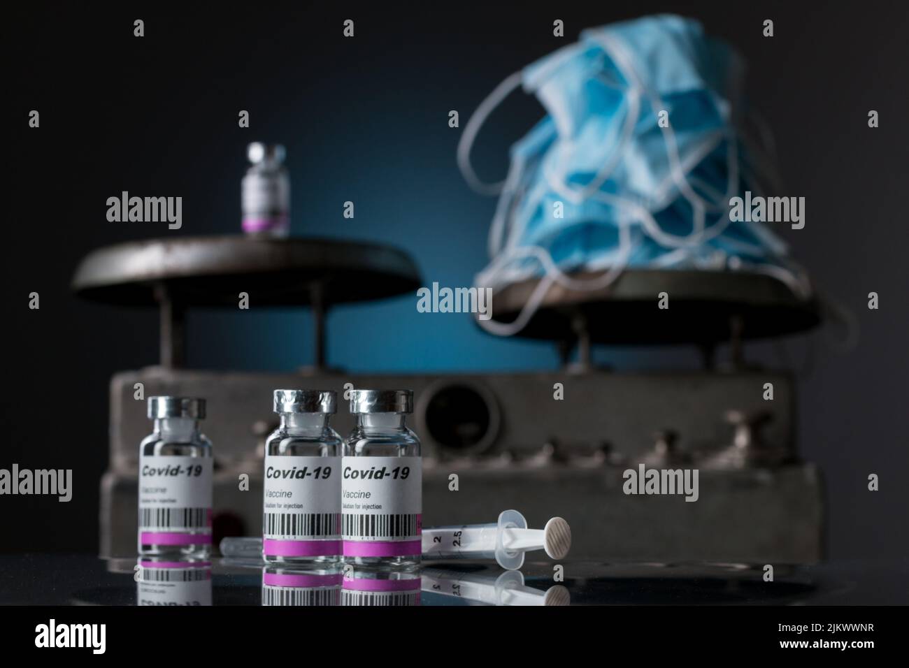 Nahaufnahme von vier ANTICOVID-Impfstofffläschchen mit Spritze und OP-Masken. Im Hintergrund eine alte Waage Stockfoto