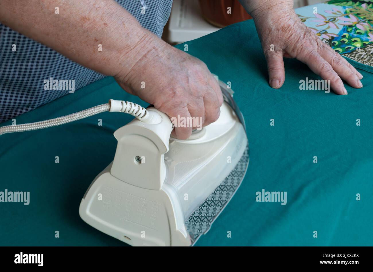 Ältere Dame beim Bügeln zu Hause. Nahaufnahme der Hände der älteren Dame beim Bügeln der Kleidung. Häusliche Arbeit mit Glück, selektiver Fokus gemacht. Stockfoto