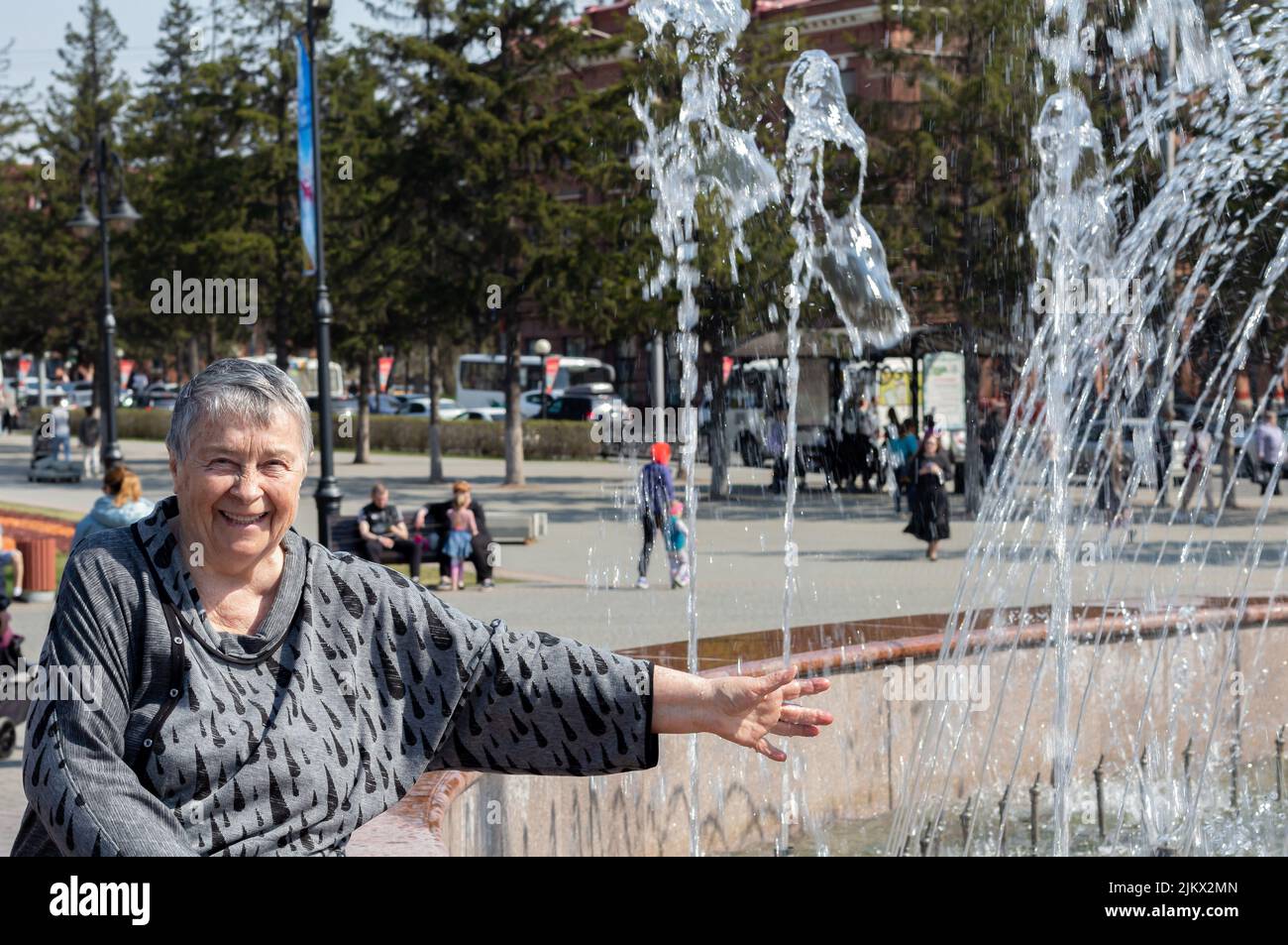 Entspannte und lachende ältere Frau, die am Brunnen sitzt, durch Düsen eines funktionierenden Brunnens mit grünen Bäumen und Sträuchern im Hintergrund gesehen, enj Stockfoto