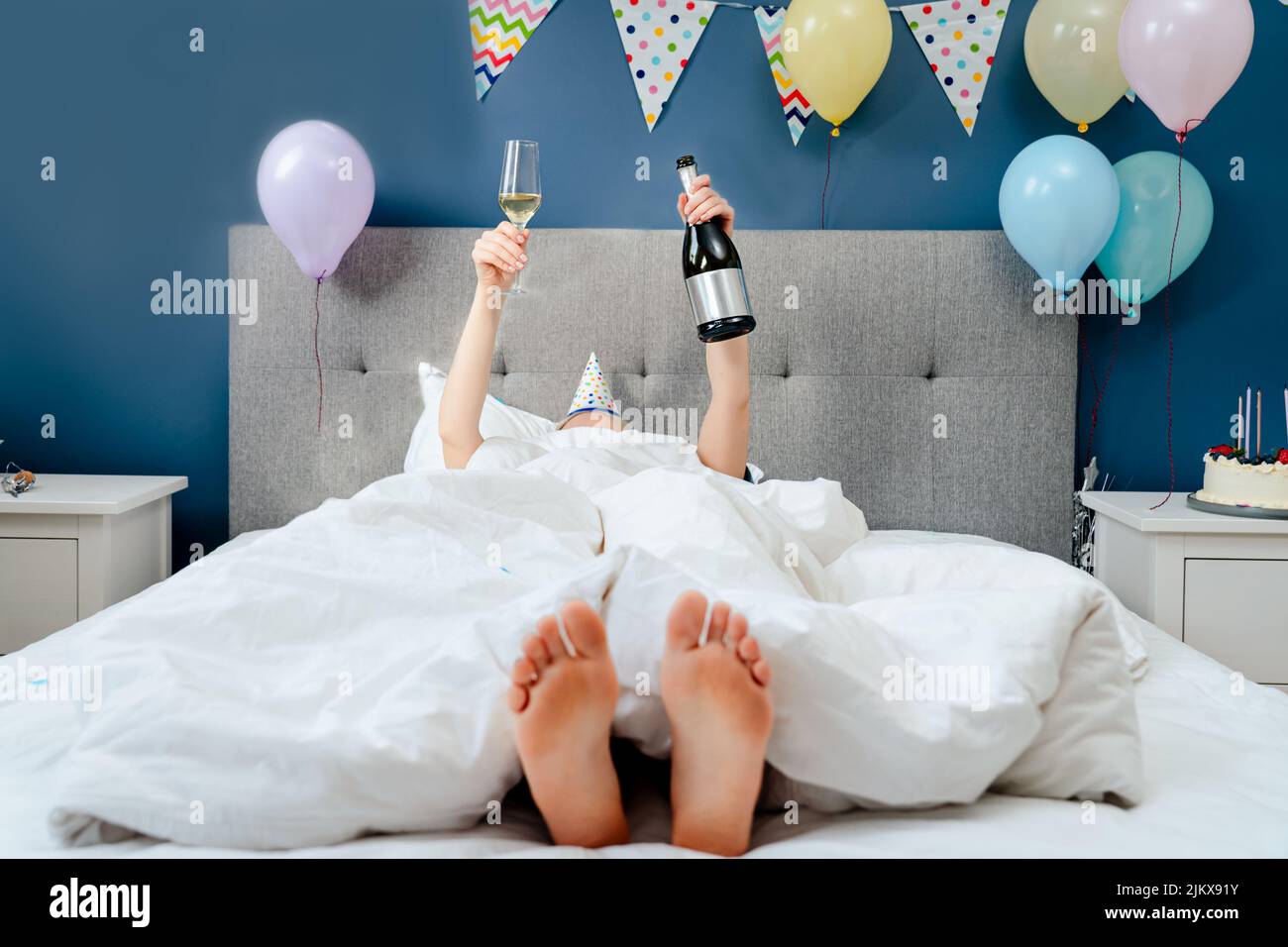 Frau liegt auf dem Bett mit erhobenen Händen halten Flasche und Glas Champagner in dekorierten Schlafzimmer.Pläne für Geburtstag. Zeit für sich selbst. Feiern Stockfoto
