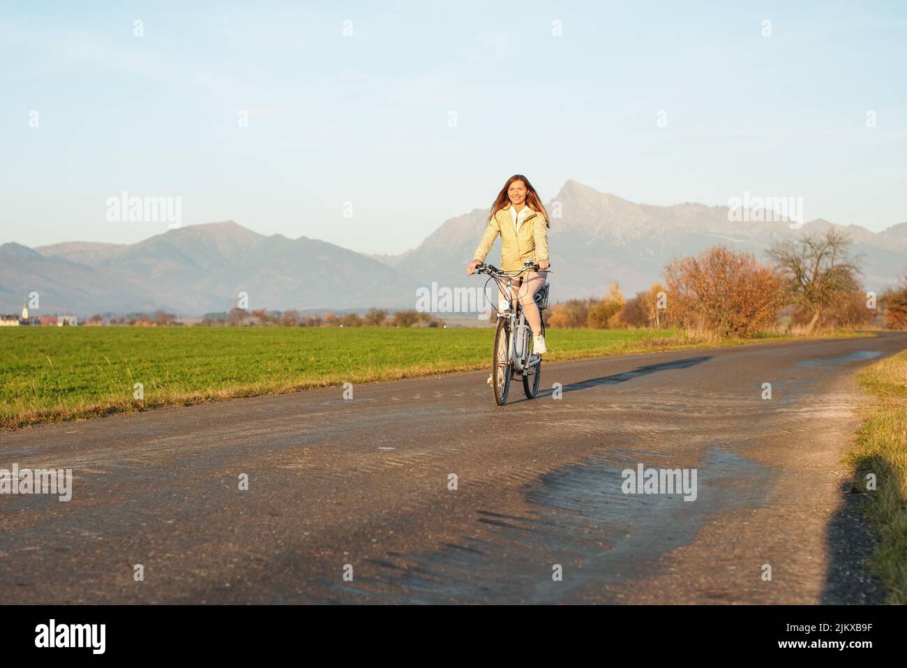 Junge Frau in Jacke fährt mit dem Fahrrad über die Landstraße, die Nachmittagssonne scheint am Berg Krivan - slowakisches Symbol - Hintergrund Stockfoto