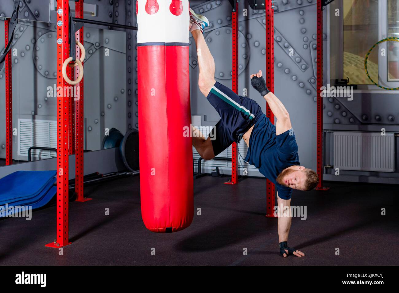 Der junge MMA-Kämpfer im invertierten Zustand liefert einen starken Straight Kick auf die rote Boxbirne in den Bereich des angeblichen Gegners. Trainingsprozess im Fitnessstudio. Stockfoto