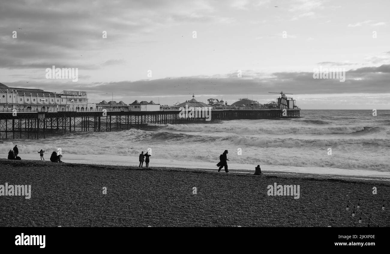 Ein Blick in Graustufen auf die Küste von Brighton und ein alter Pier mit Menschen, die sich am Strand, Großbritannien, ausruhen Stockfoto