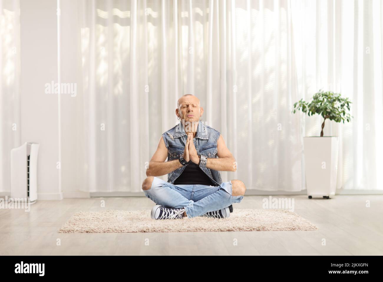 Kahlköpfiger Mann in Jeans sitzt mit gekreuzten Beinen und praktiziert Yoga zu Hause auf einem Teppich Stockfoto