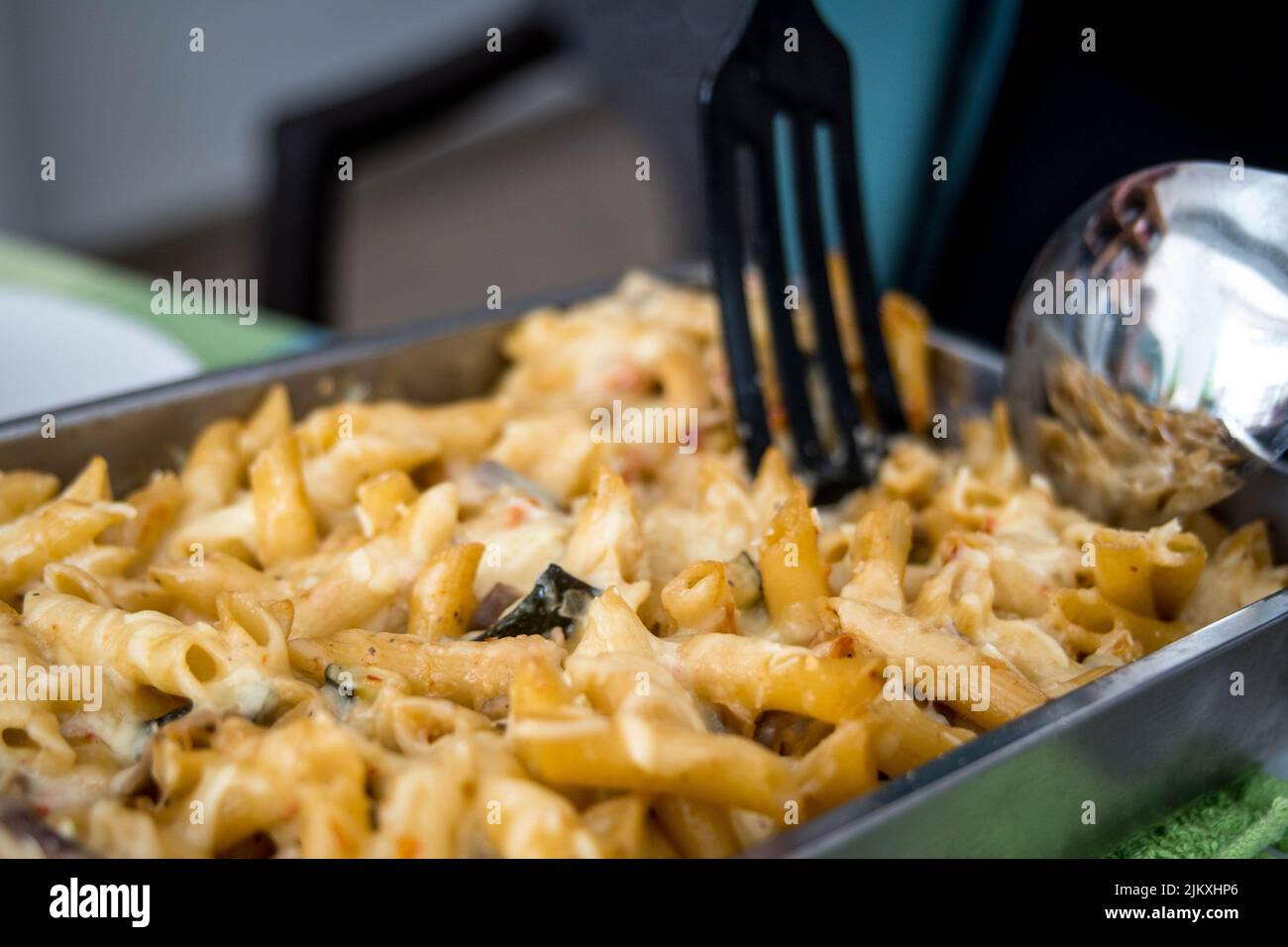 Bild von Tablett mit Macaroni Pasta mit geschmolzenem Käse und Gemüse, das während einer Mahlzeit mit Paddel und großem Löffel am Tisch serviert wird Stockfoto