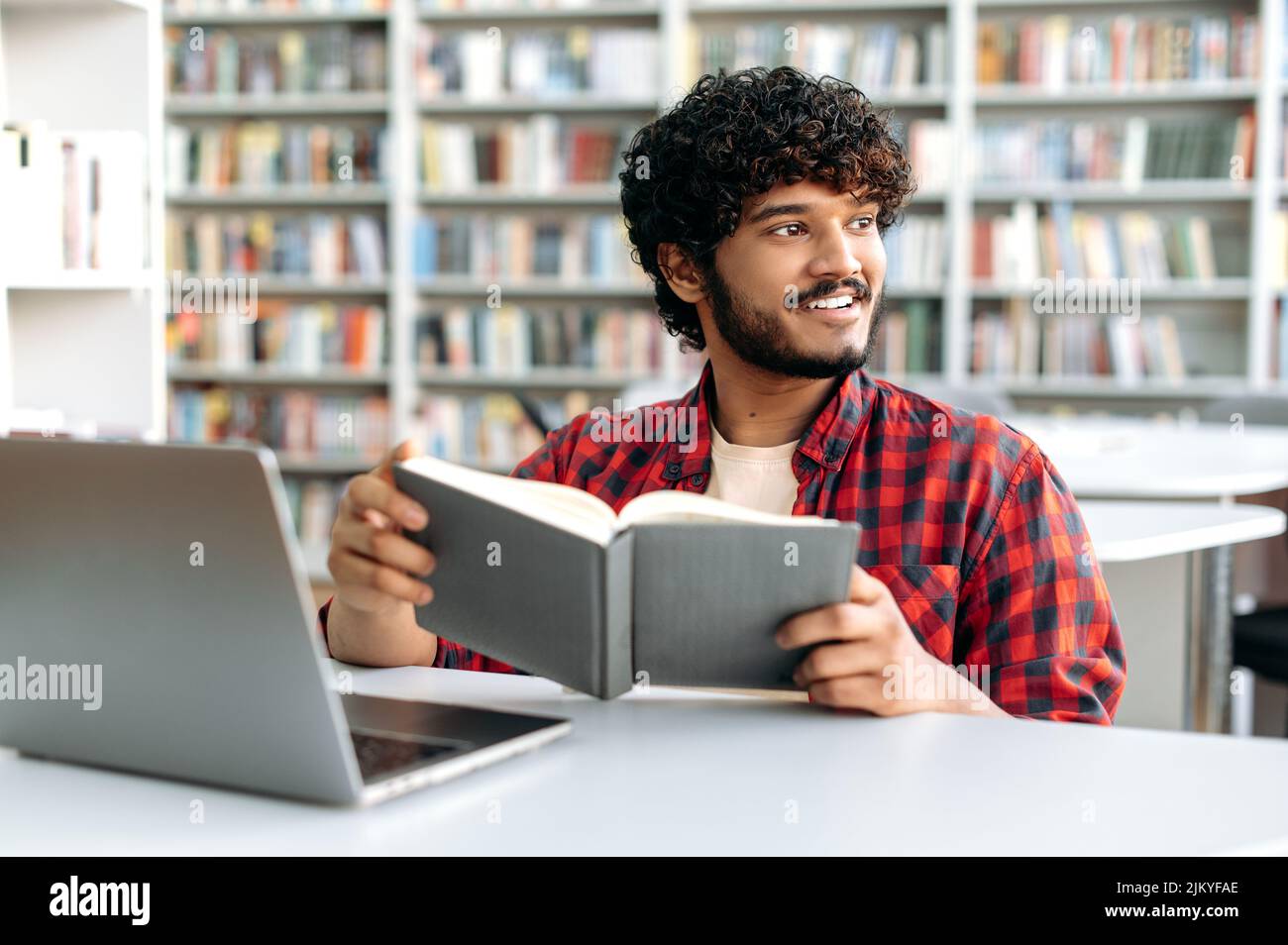 Positive intelligente arabische oder indische College-Student, sitzt am Tisch mit Laptop in der Universitätsbibliothek, Vorbereitung auf die Prüfung, Studium von Informationen, Buch halten, wegschauen, Träumen von Urlaub, Lächeln Stockfoto