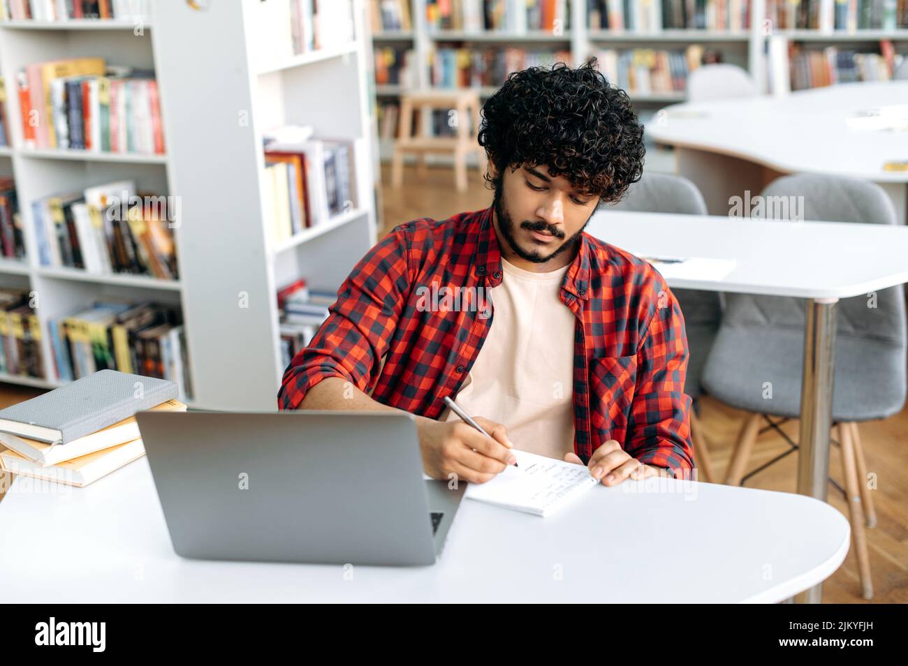 Fokussierter, kluger arabischer oder indischer Student, sitzt mit einem Laptop in der Universitätsbibliothek am Tisch, bereitet sich auf die Prüfung vor, studiert Informationen, hört Vorträge, notiert im Notizbuch, lernt Wissen Stockfoto