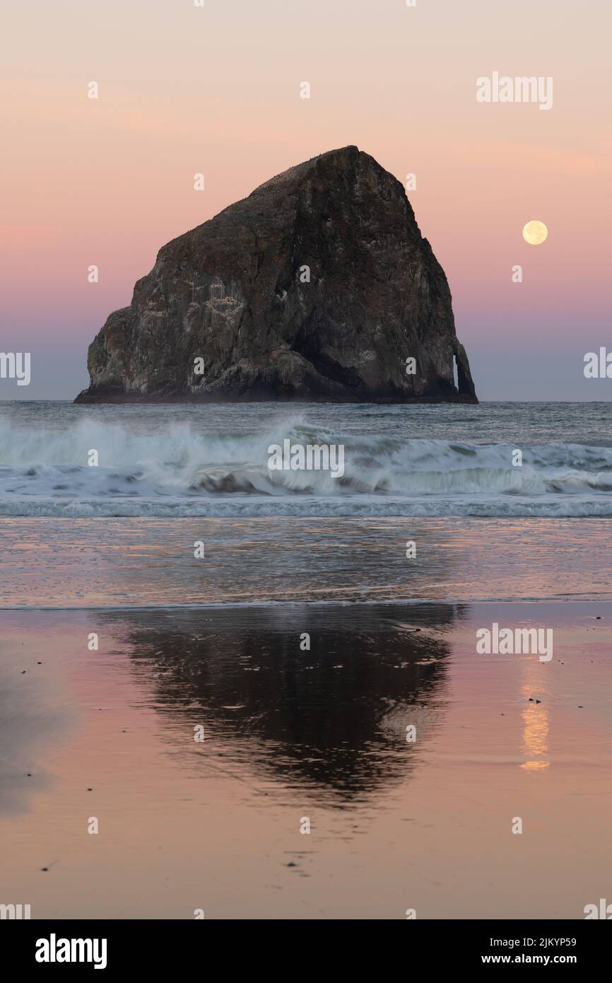 Im Morgengrauen stürzen Wellen vor dem Haystack Rock in Pacific City Oregon an Land, während der Vollmond an einem bunten Horizont untergeht. Stockfoto