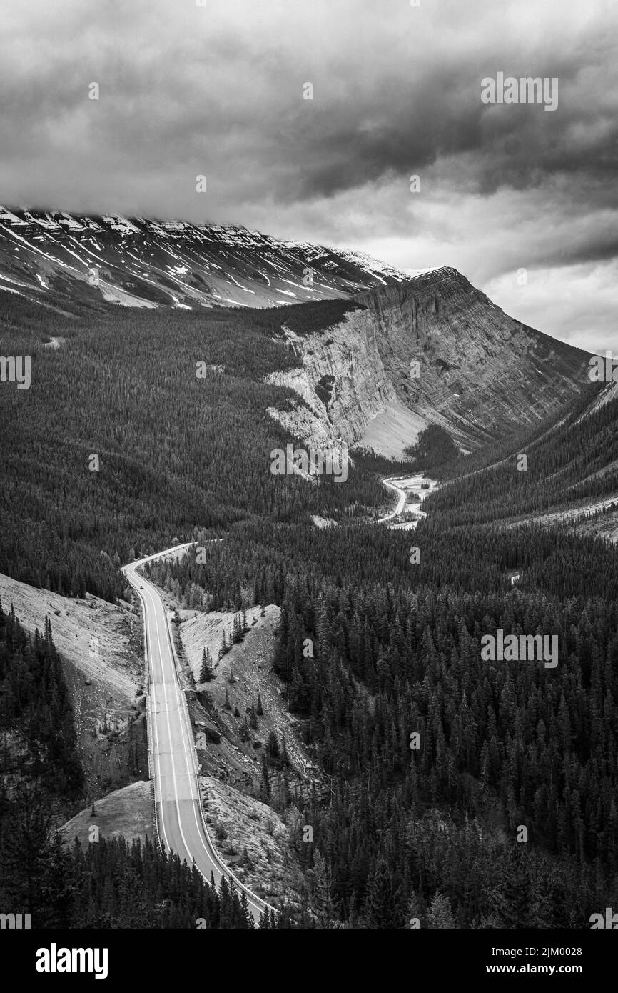 Eine vertikale Graustufenansicht einer Straße, die an einem bewölkten Tag durch Berge führt Stockfoto