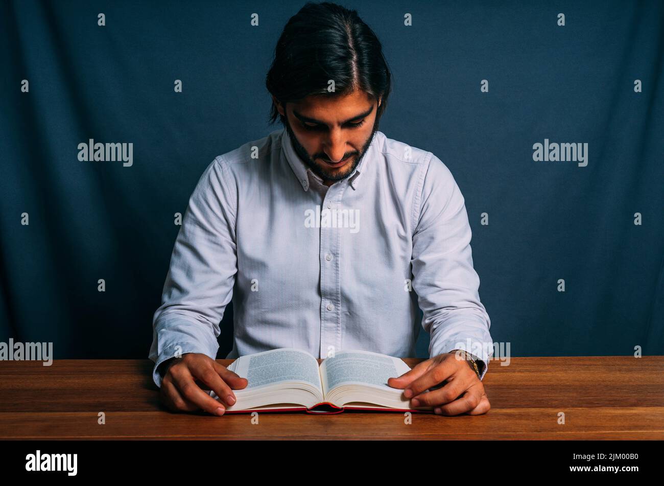 Bild eines stylischen hispanischen Mannes im gestreiften Hemd, das studiert, konzentriert ein großes Buch, das an einem Holztisch sitzt. Konzept des harten Studiums Stockfoto