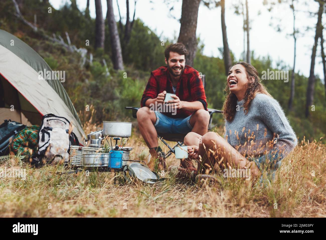 Glückliches junges Paar, das vor ihrem Zelt lachend sitzt. Fröhliches junges Paar, das sich auf einem Campingurlaub amüsieren kann. Stockfoto