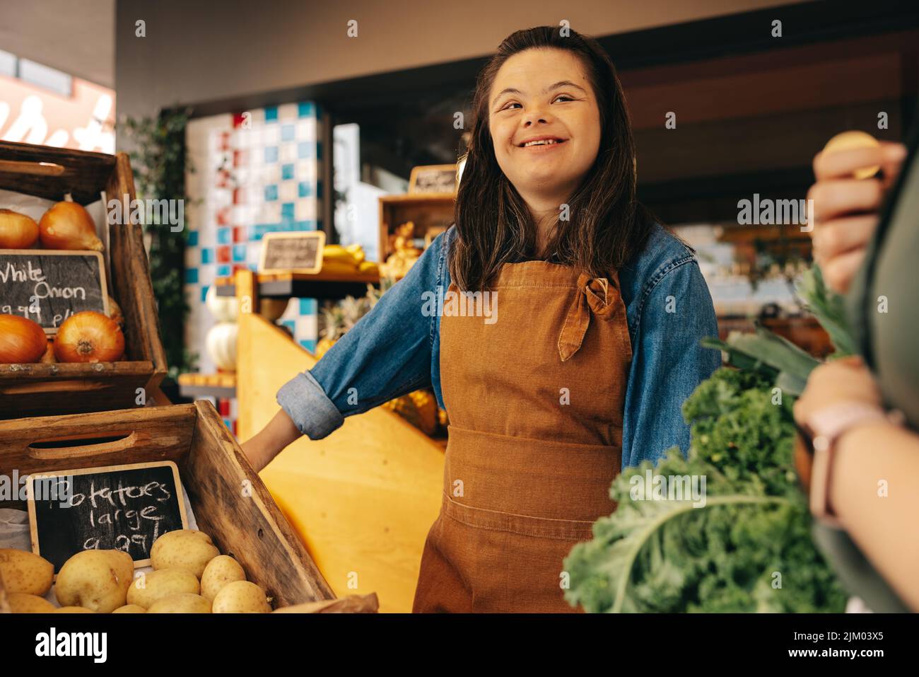 Glückliche Frau mit Down-Syndrom, die einem Kunden in der Gemüseabteilung eines Lebensmittelladens hilft. Freundliche Frau mit einer intellektuellen Behinderung arbeitet Stockfoto