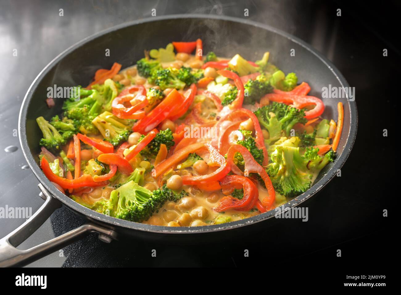 Dampfendes Gemüsekry mit Kichererbsen, Brokkoli und rotem Paprika in Kokosmilch in einer Pfanne auf einem schwarzen Herd, gesunde Mahlzeiten für einen veganen Low Stockfoto