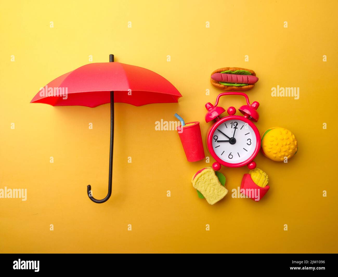 Roter Regenschirm mit Essen und Wecker auf gelbem Hintergrund. Das Konzept der Lebensmittelsicherheit. Stockfoto