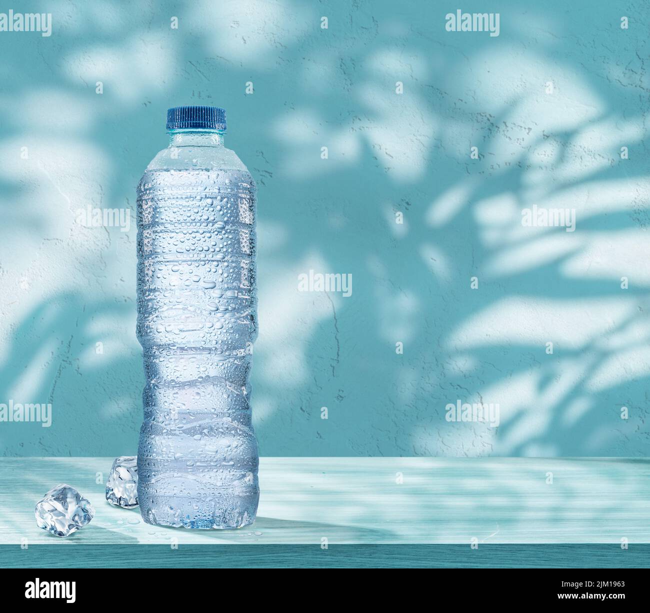 Plastikflasche mit gekühltem Wasser mit Kondensationstropfen und zwei Eiswürfeln auf dem Tisch. Blauer Hintergrund. Stockfoto
