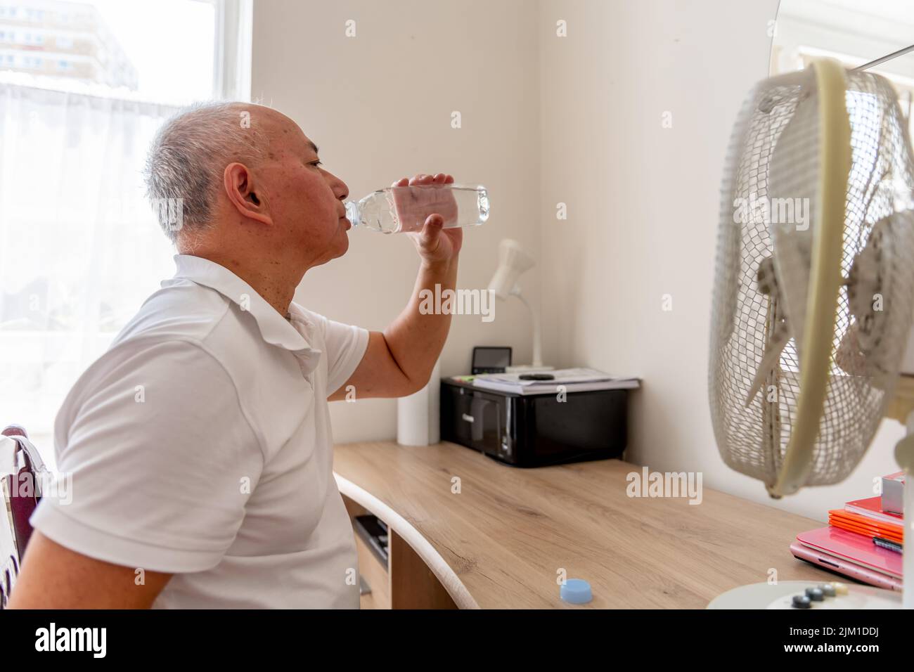 Ein älterer Mann zu Hause, der an einem rekordverdächtigen heißen Sommertag neben einem elektrischen Ventilator Wasser trinkt und versucht, Dehydrierung und Hitzschlag zu vermeiden. Stockfoto