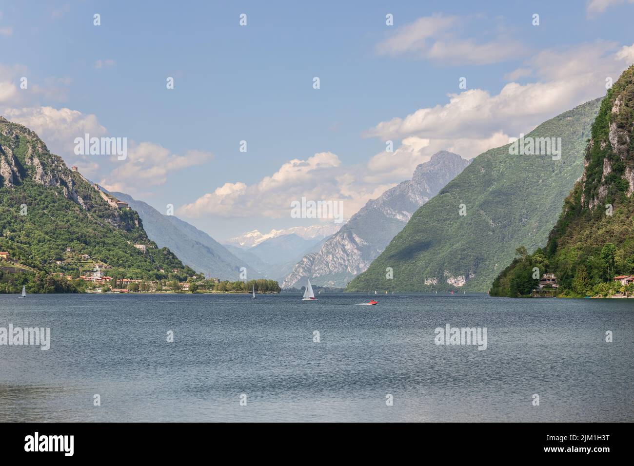 Segelboote und ein rotes Motorboot kreuzen den welligen Idrosee zwischen bewaldeten Kalksteinbergen in der Lombardei, Italien, unter einem blauen Frühlingshimmel. Stockfoto