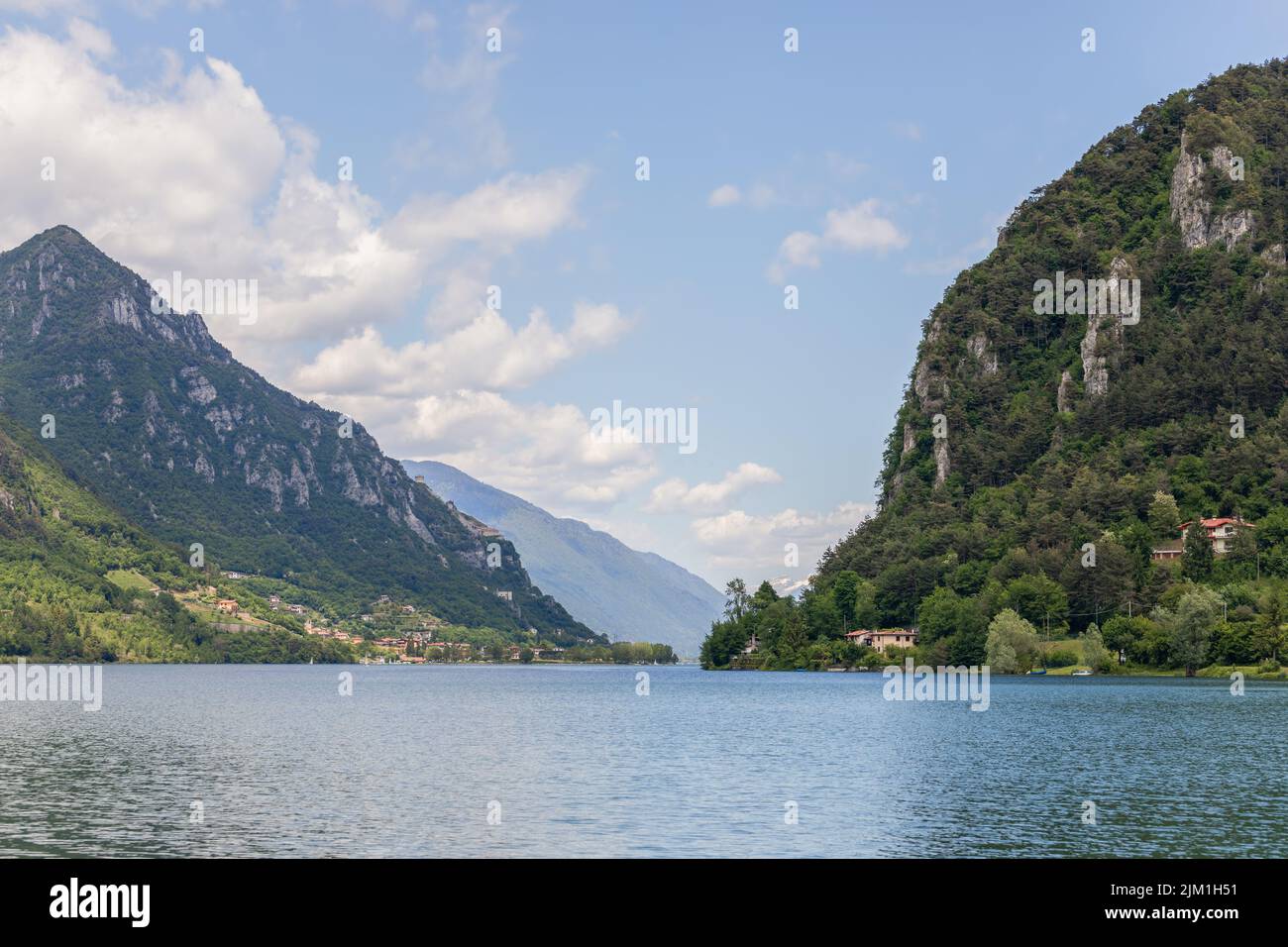 Der Idrosee, Lombardei, Italien, verengt sich zwischen bewaldeten Klippen, mit kleinen Uferhäusern und fernen blauen Bergrücken unter vereinzelten Wolken. Stockfoto