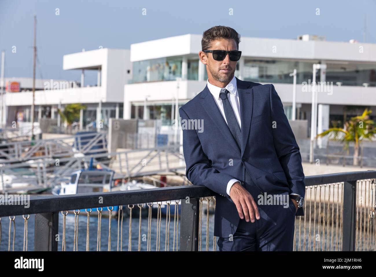 Ernsthafter Geschäftsmann in Anzug und Sonnenbrille, der sich am Brückengeländer lehnt und am sonnigen Tag im Stadthafen auf ein modernes Gebäude blickt Stockfoto