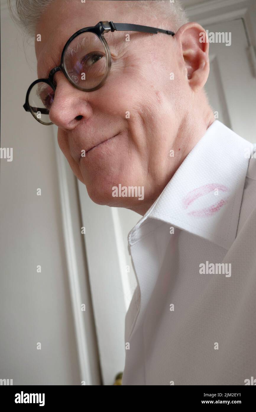 Ein älterer Mann schmunkt und zeigt einen Lippenstift-Flecken auf seinem weißen Hemdkragen Stockfoto