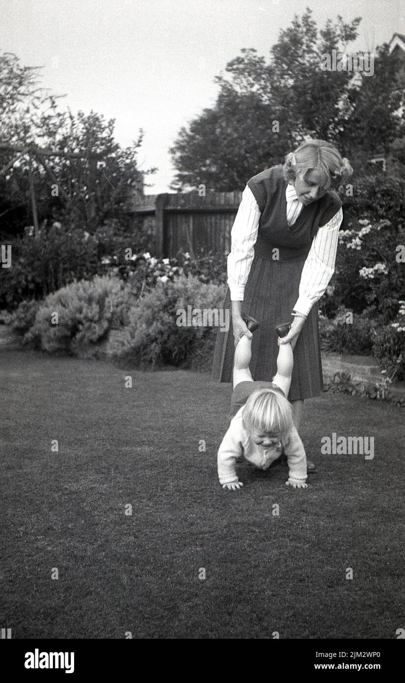 1960s, historischer, kindlicher Spaß draußen in einem Garten, ein kleines Mädchen, das auf einem Rasen mit dem Schubkarren unterwegs ist, Mutter mit ihren Unterschenkeln und Knöcheln, England, Großbritannien Stockfoto