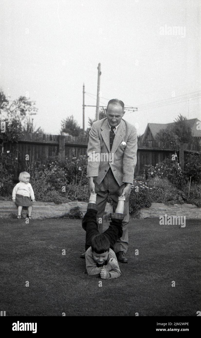 1960s, historisch, ein kleiner Junge, der sich draußen in einem Garten beim Laufen mit dem Schubkarren amüsieren kann, wobei sein Onkel seine Unterschenkel in der Hand hält und ankes, England, Großbritannien Stockfoto
