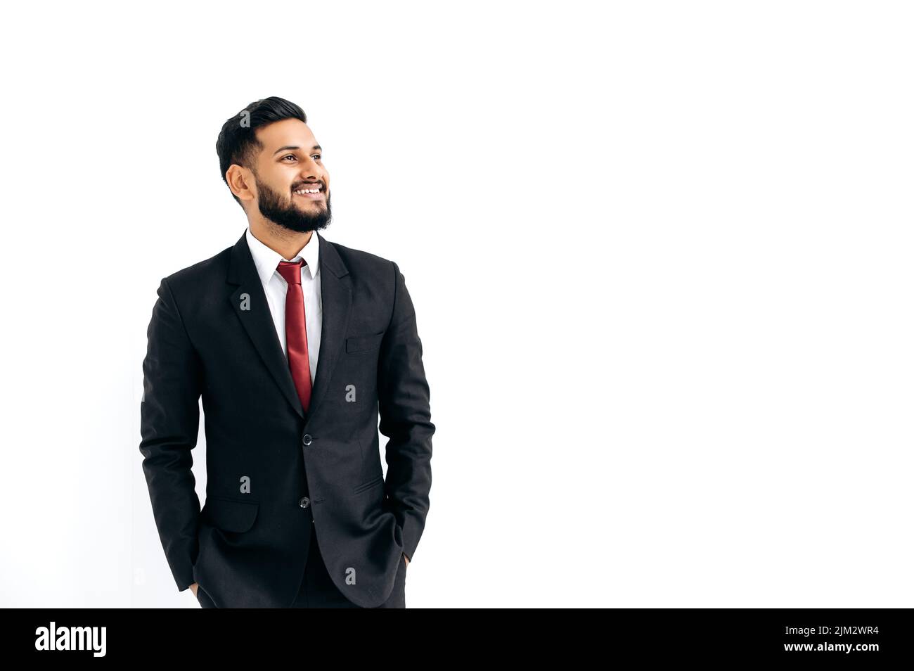Platz zum Kopieren Ihrer Präsentation. Freundlicher, eleganter, einflussreicher arabischer oder indischer Mann in formellem Anzug, stehend auf isoliertem weißem Hintergrund, wegblickend, denkend, planend, träumend, Lächelt fröhlich Stockfoto