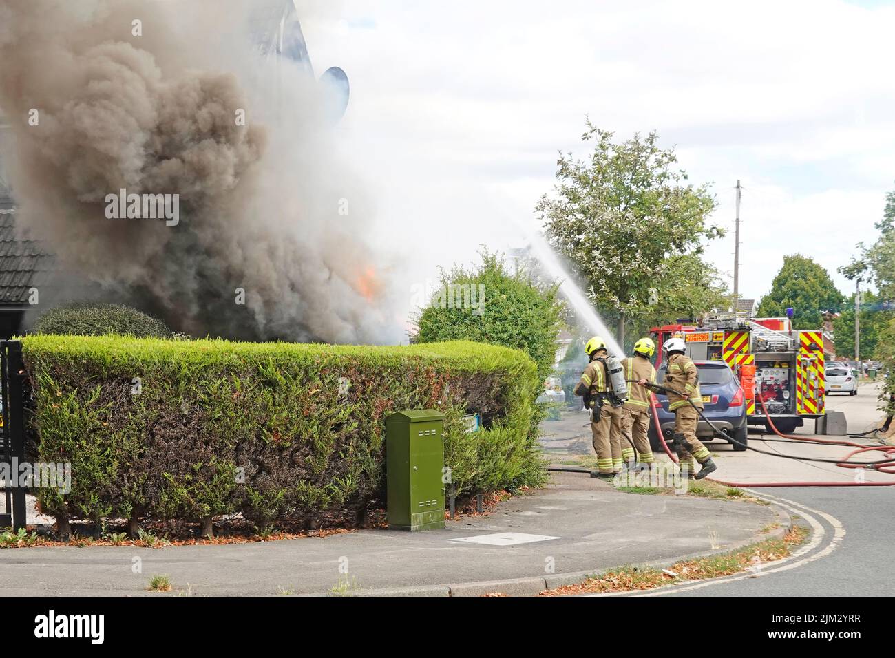 Rauch und Flammen wehend als Gruppe von Feuerwehrmänner zielen Wasserstrahl an Hausfront und Seitenwänden Essex Fire & Rescue Tender Wohnstraße Essex England UK Stockfoto