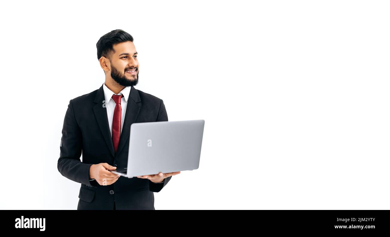 Eleganter, erfolgreicher indischer oder arabischer junger Mann im Business-Anzug, männlicher Manager, der einen offenen Laptop in den Händen hält, zur Seite schaut, auf isolierten weißen Hintergrund steht und freundlich lächelt. Copy-Space-Konzept Stockfoto