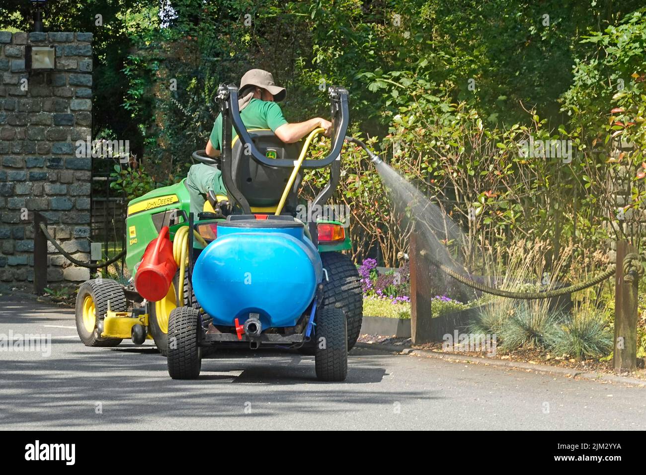 Gärtner, der hoch oben auf dem John Deere Minitraktor sitzt und einen blauen Mini-Pumpwassertanker schleppt, der Blumen im Landgut des Hotels England versprüht Stockfoto