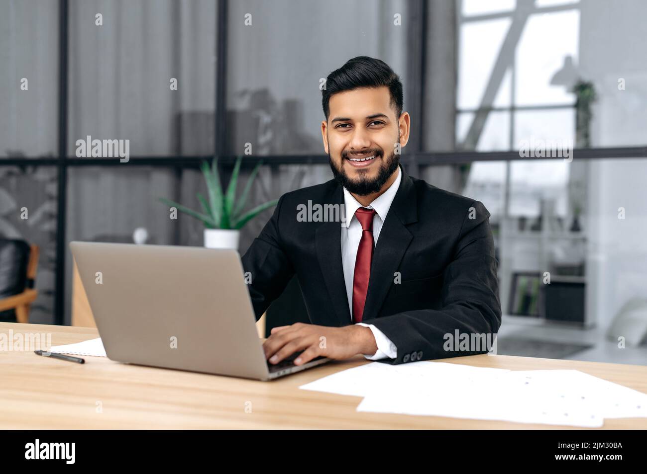 Porträt eines gutaussehenden, selbstbewussten arabischen oder indischen erfolgreichen Geschäftsmannes, Unternehmers, Rechtsanwalts, in einem formellen Anzug, sitzen am Schreibtisch mit Laptop in einem modernen kreativen Büro, blickt auf die Kamera, lächeln freundlich Stockfoto