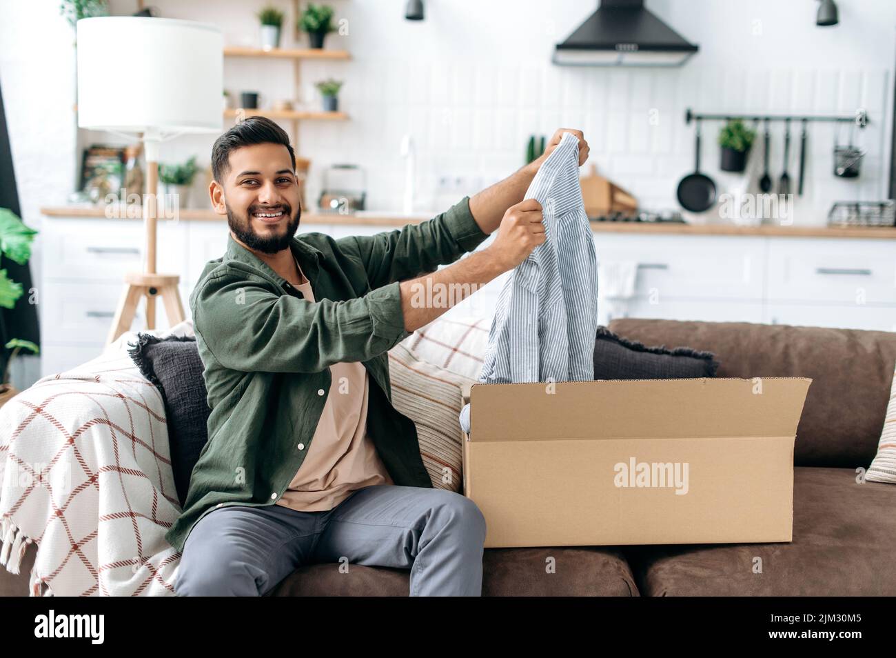 Online-Shopping-Konzept. Fröhlicher arabischer oder indischer junger Mann, der sein Paket auspackt, glücklich darüber, eine lange erwartete Bestellung zu bekommen. Gemischtes Rennen moderner Kerl Einkaufen in Internet-Shops, neue Kleidung online kaufen Stockfoto