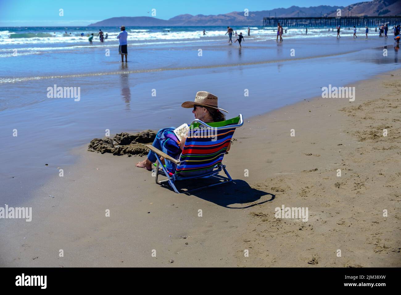 Lady in Sonnenhut sitzt auf einem Stuhl am Ufer und liest ein Buch am Pismo Beach, Kalifornien, mit Bergen und Pismo Pier im Hintergrund. Stockfoto